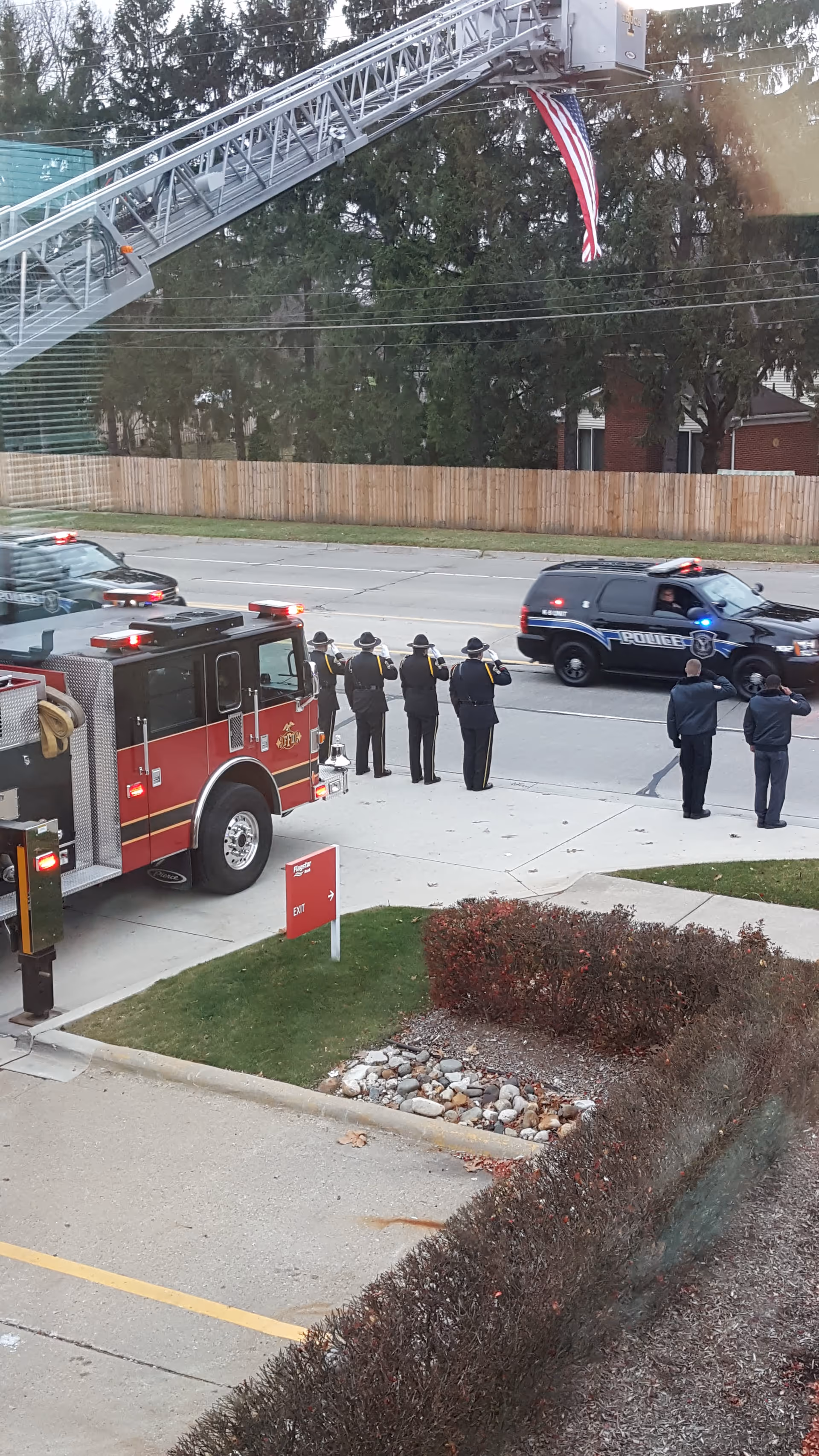 Emergency responders stand by a fire truck and police SUV on a street; flag on ladder.