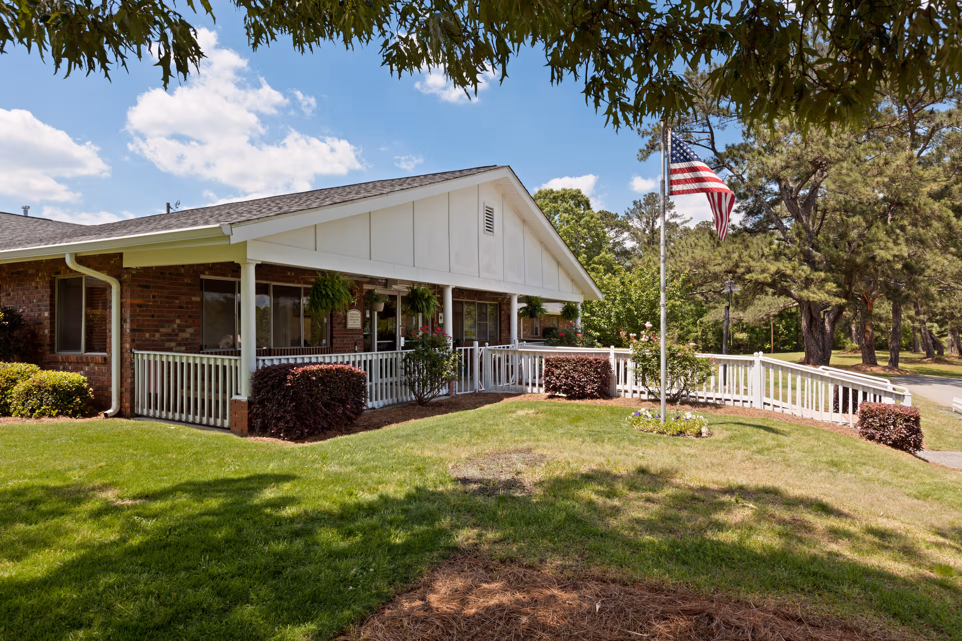 Front exterior of a senior living facility with white porch railing, flagpole, and trees.
