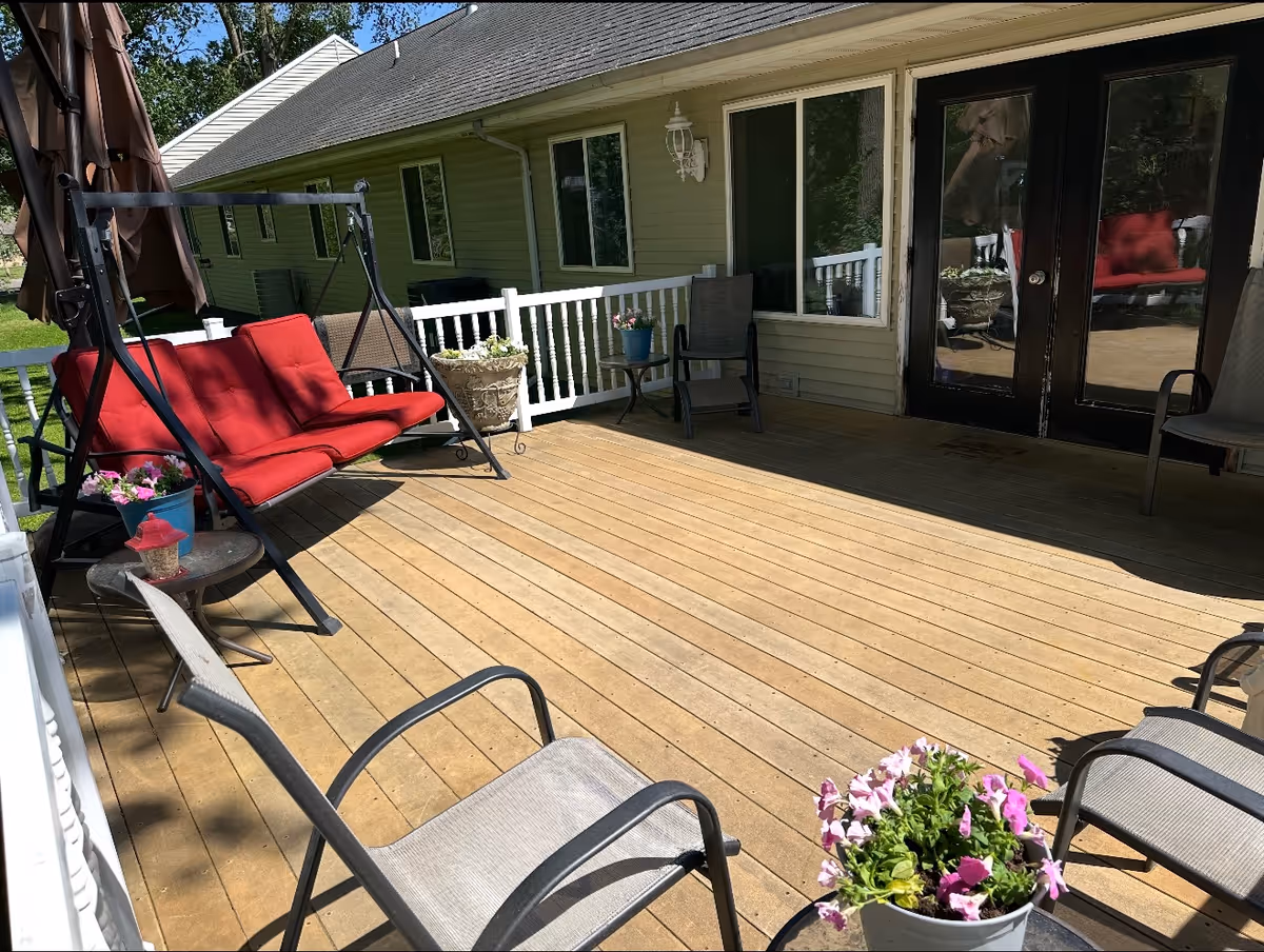 Sunny wooden deck with a red cushioned swing, patio chairs, potted flowers, and glass doors into the building.