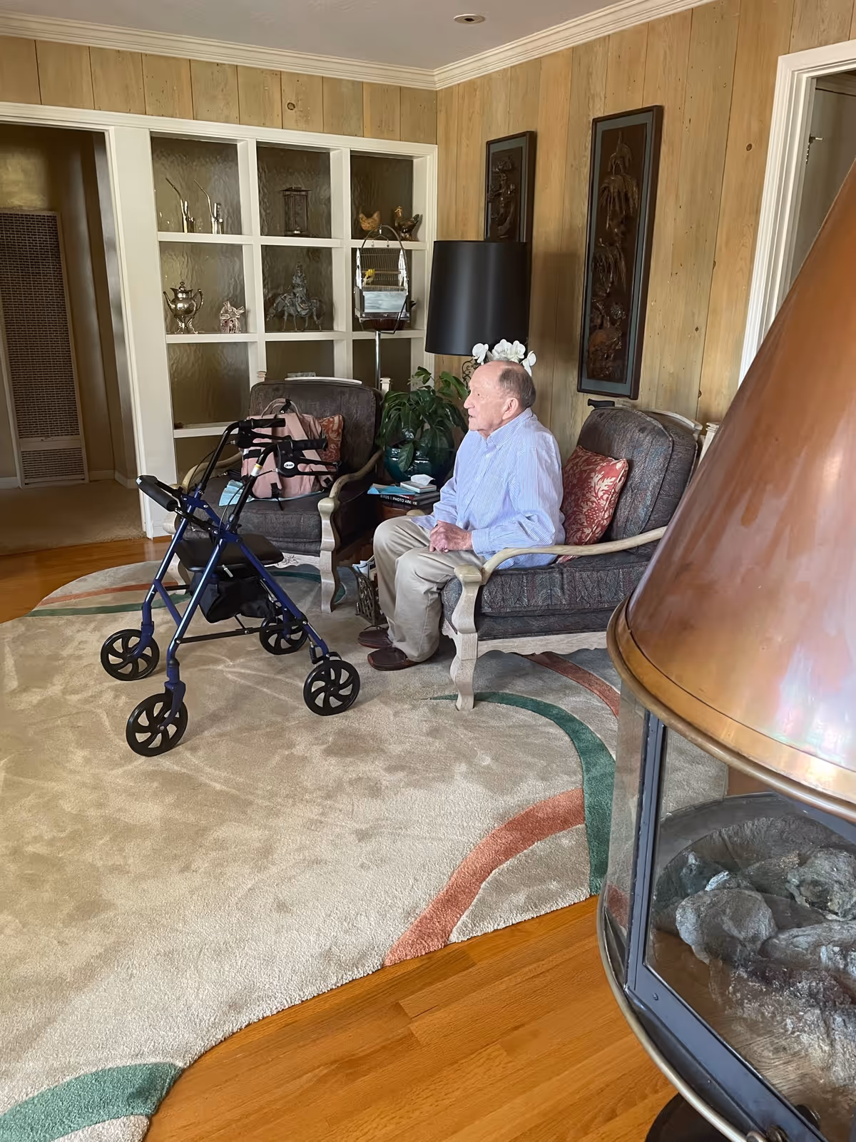 An elderly man sits in a living room with a walker nearby and decorative shelves.