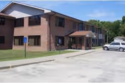 Two-story brick senior living building with a covered entrance, sidewalk, and a parking lot with a white SUV.