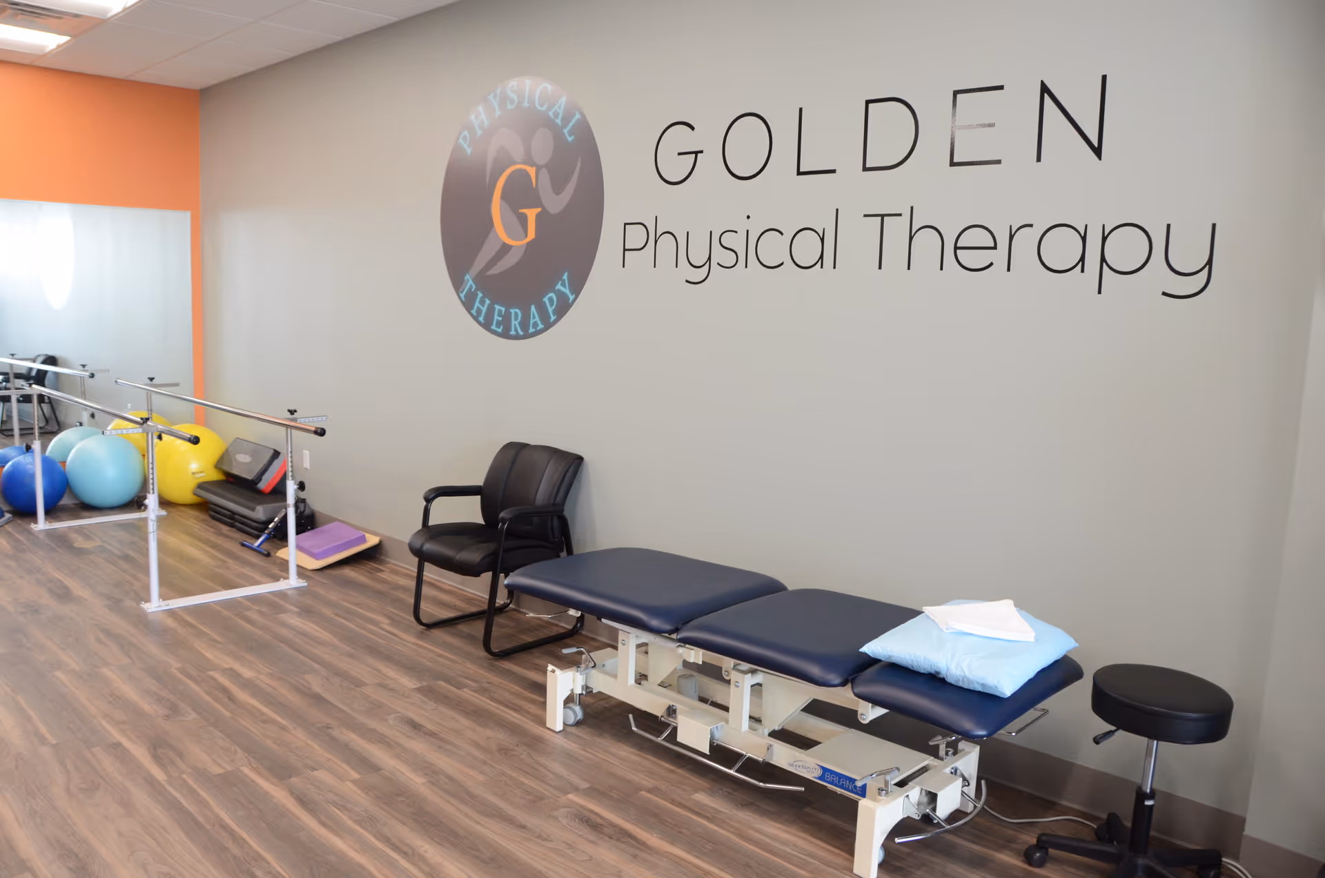 Physical therapy treatment room with a treatment table, chairs, balls, and parallel bars.