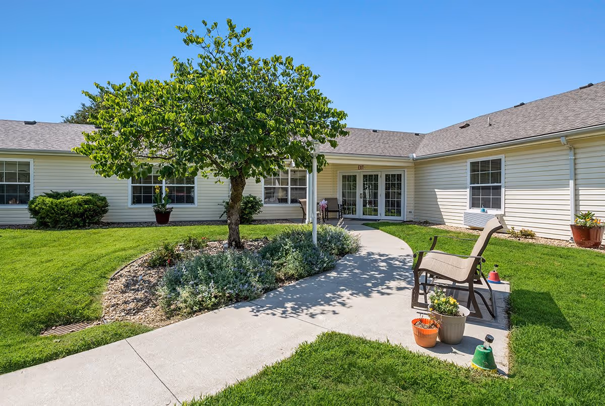 Courtyard and entrance of a single-story assisted living building with a tree, walkway, lawn, and outdoor seating.