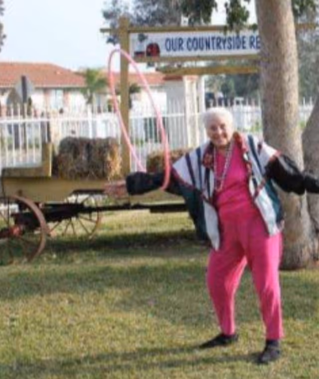 An elderly woman wearing a colorful jacket and pink pants is playing with a pink hula hoop outdoors on a grassy area. Behind her, there is a wooden wagon with hay bales and a sign that reads 'Our Countryside Resort'. Trees and a white fence are visible in the background.