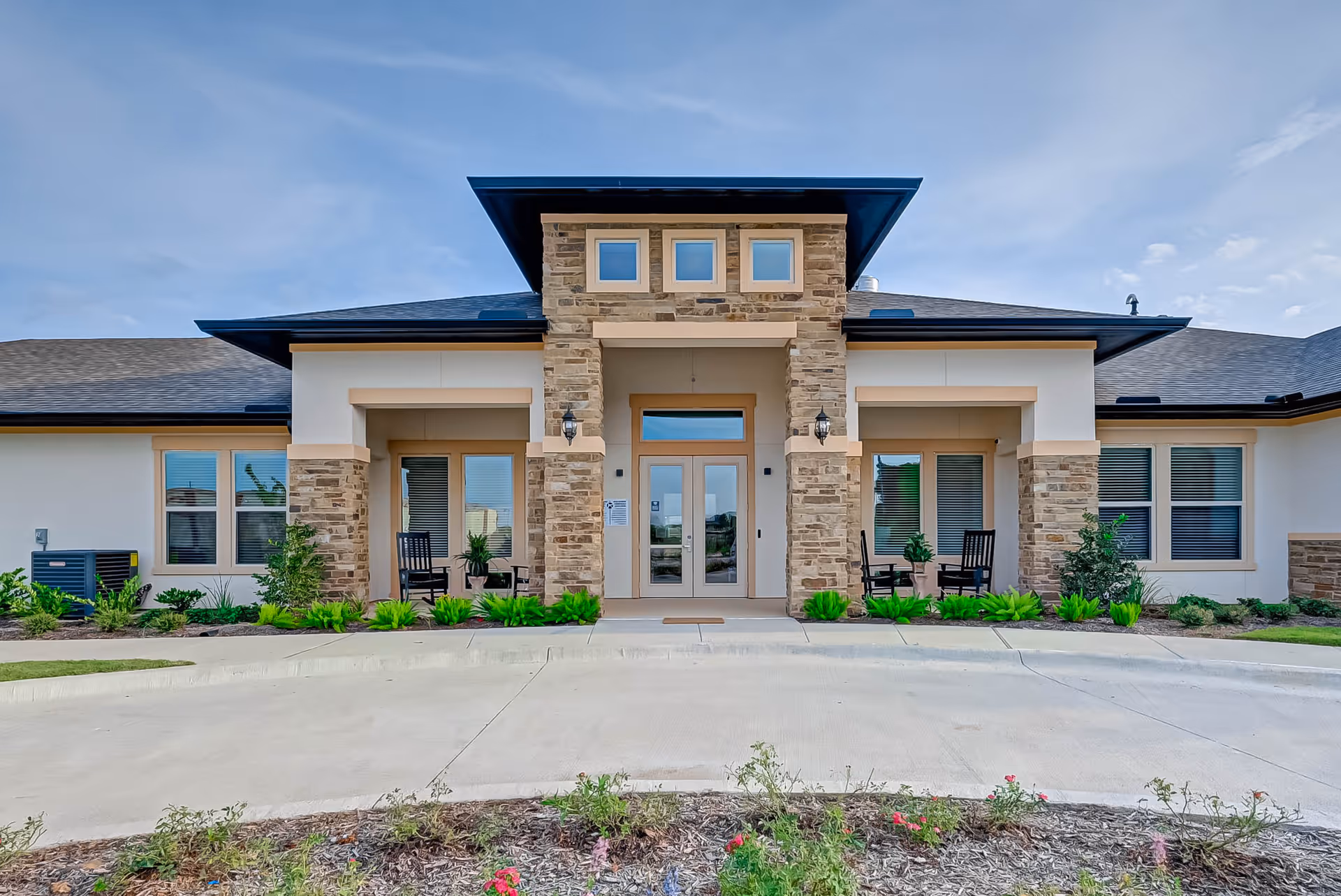 Front exterior view of a single-story assisted living and memory care facility with stone and beige facade, large glass double doors at the entrance, two seating areas with black chairs and small tables on either side, and landscaped greenery in front.