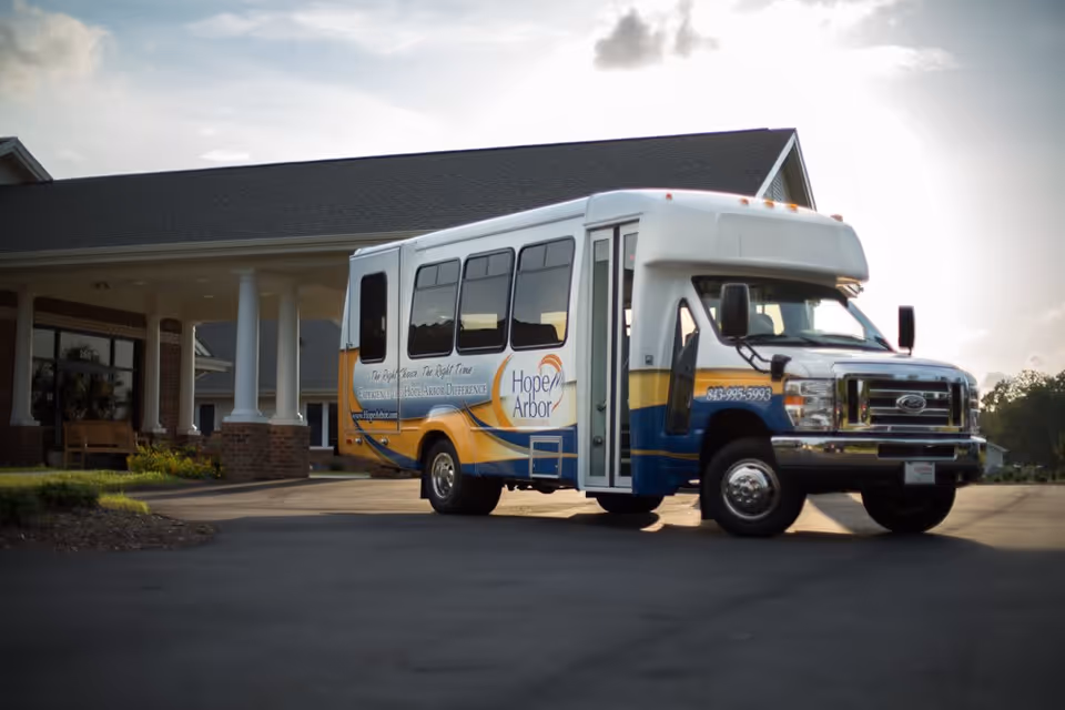 A white and blue shuttle bus with yellow accents parked in front of a building with white columns and a covered entrance. The bus has the logo and name 'Hope Arbor' along with contact information and a website on its side.
