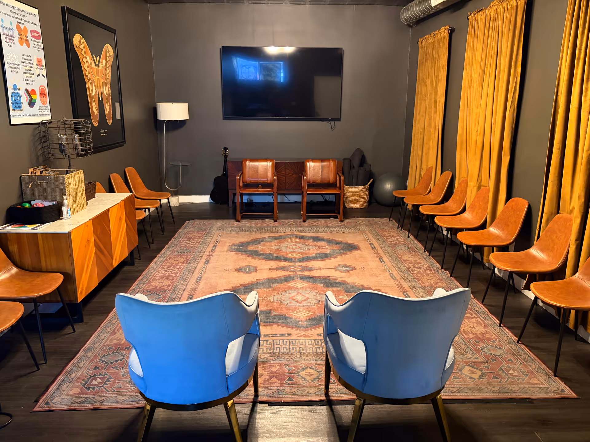 A lounge with blue armchairs, wooden wall chairs, and a TV on a dark wall.