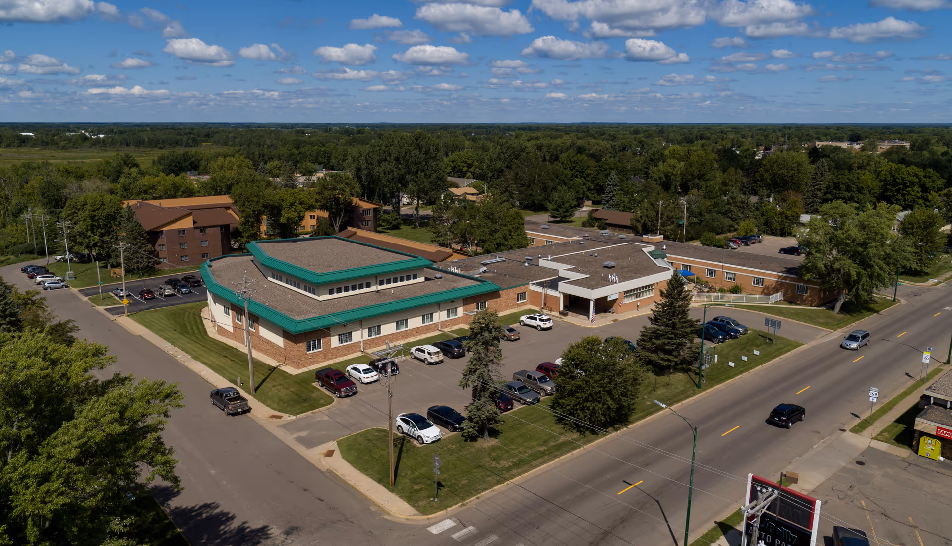 Aerial view of Aitkin Health Services building and parking lots surrounded by trees and streets under a partly cloudy sky.