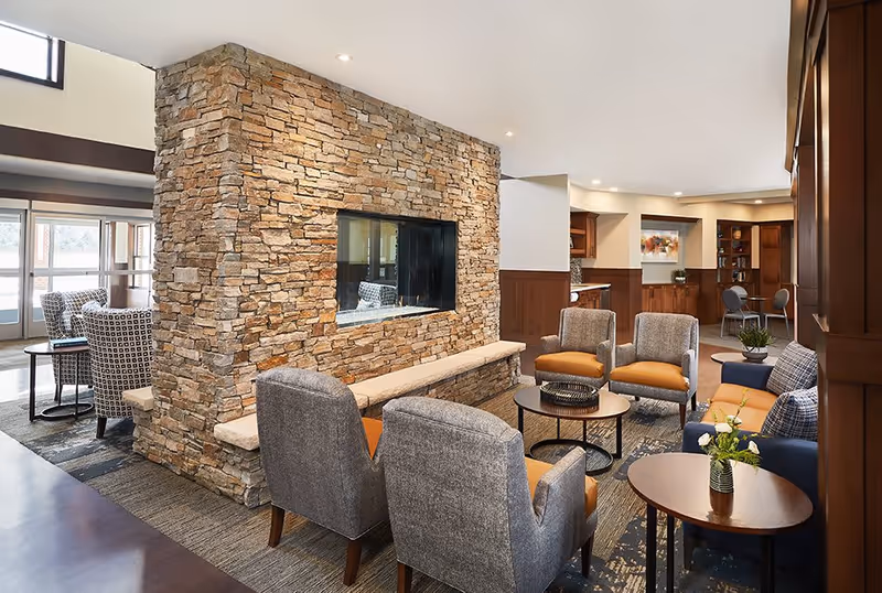 A cozy senior living facility common area featuring a stone fireplace with a built-in bench, surrounded by comfortable armchairs and sofas in gray and orange tones. The room has wood paneling, a patterned carpet, and small tables with decorative plants. In the background, there is a small dining or meeting area with chairs and a colorful painting on the wall.
