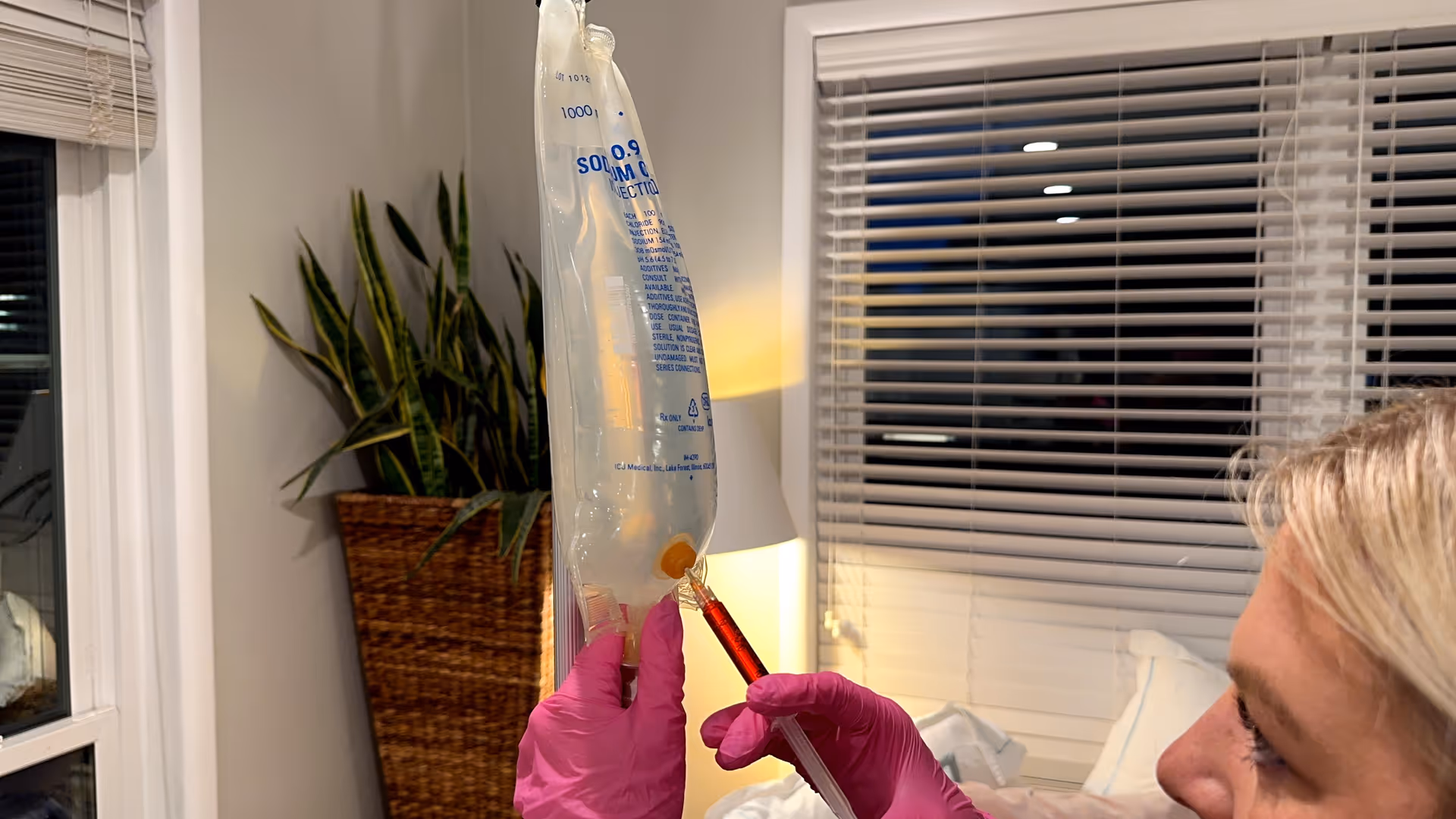 A caregiver in pink gloves administers an IV bag at a bedside in a bedroom.