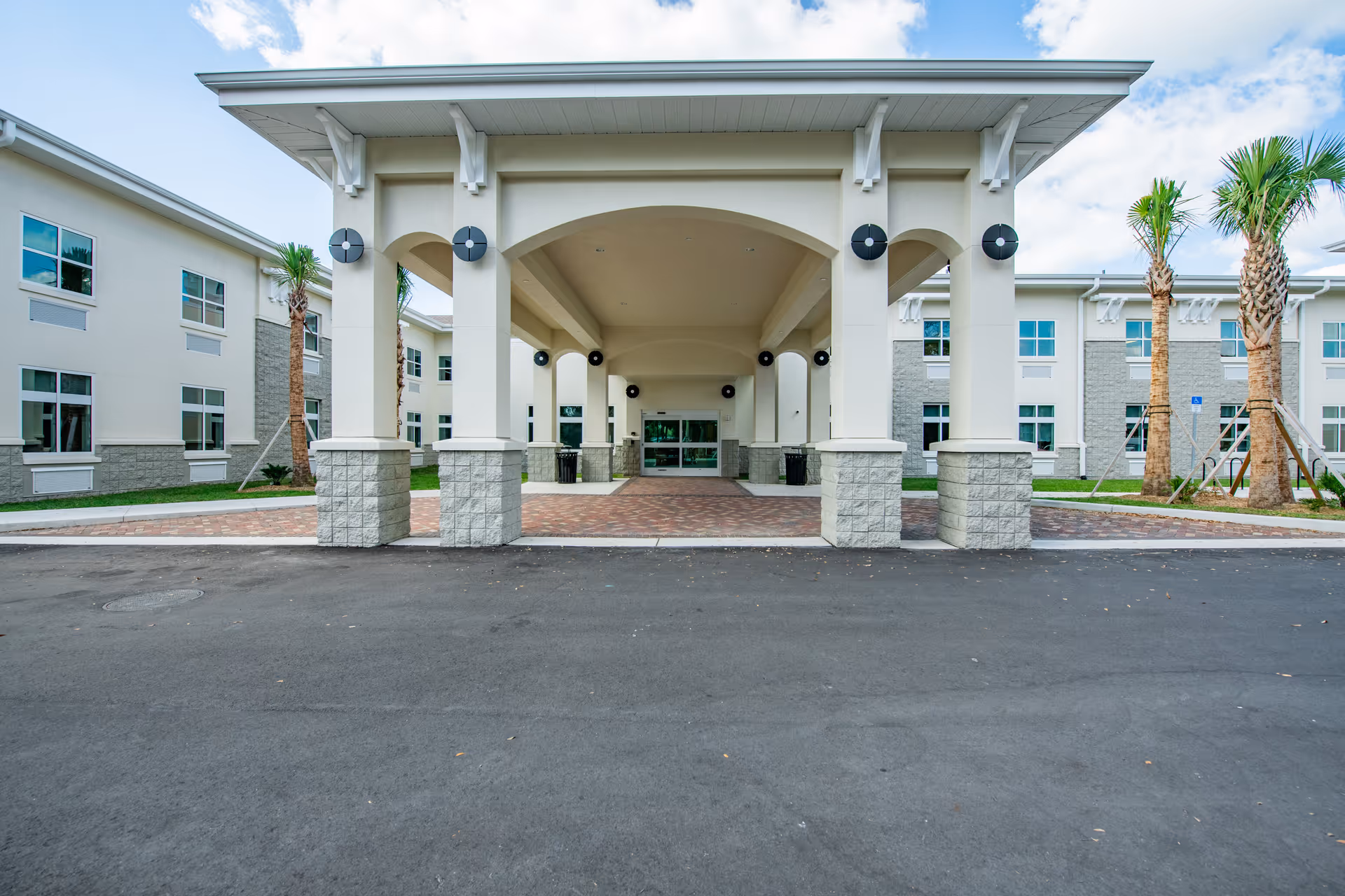 Front exterior view of Pines of Sarasota Health Care, LLC showing a covered entrance with large white columns and a paved driveway. The building is two stories with multiple windows and palm trees on either side of the entrance under a partly cloudy sky.