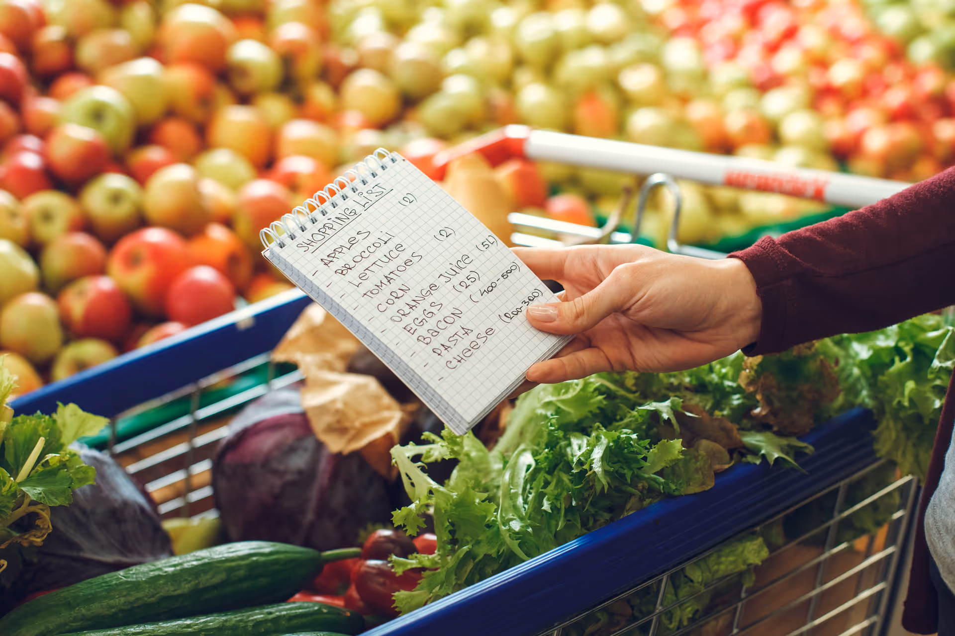 A hand holds a small shopping list beside fresh produce in a grocery store.