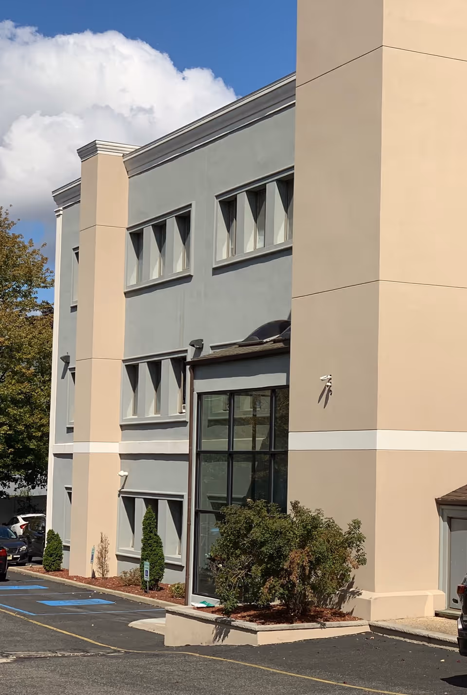 Modern senior living facility exterior with glass entry, beige and gray facade, and parking lot.