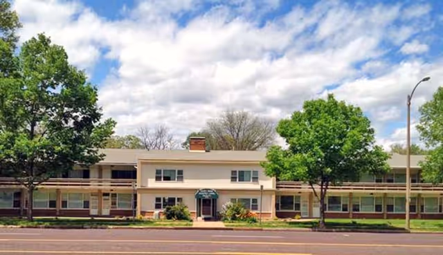 Front exterior of a two-story assisted living building with a central entrance awning, trees on either side, and a street in the foreground.