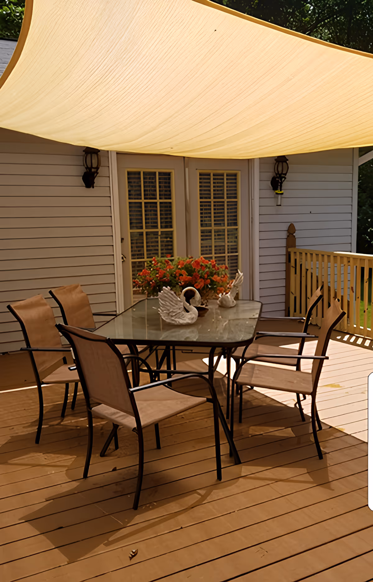 Shaded backyard deck with a glass-top table, six chairs, swan ornaments and a planter of flowers in front of French doors.