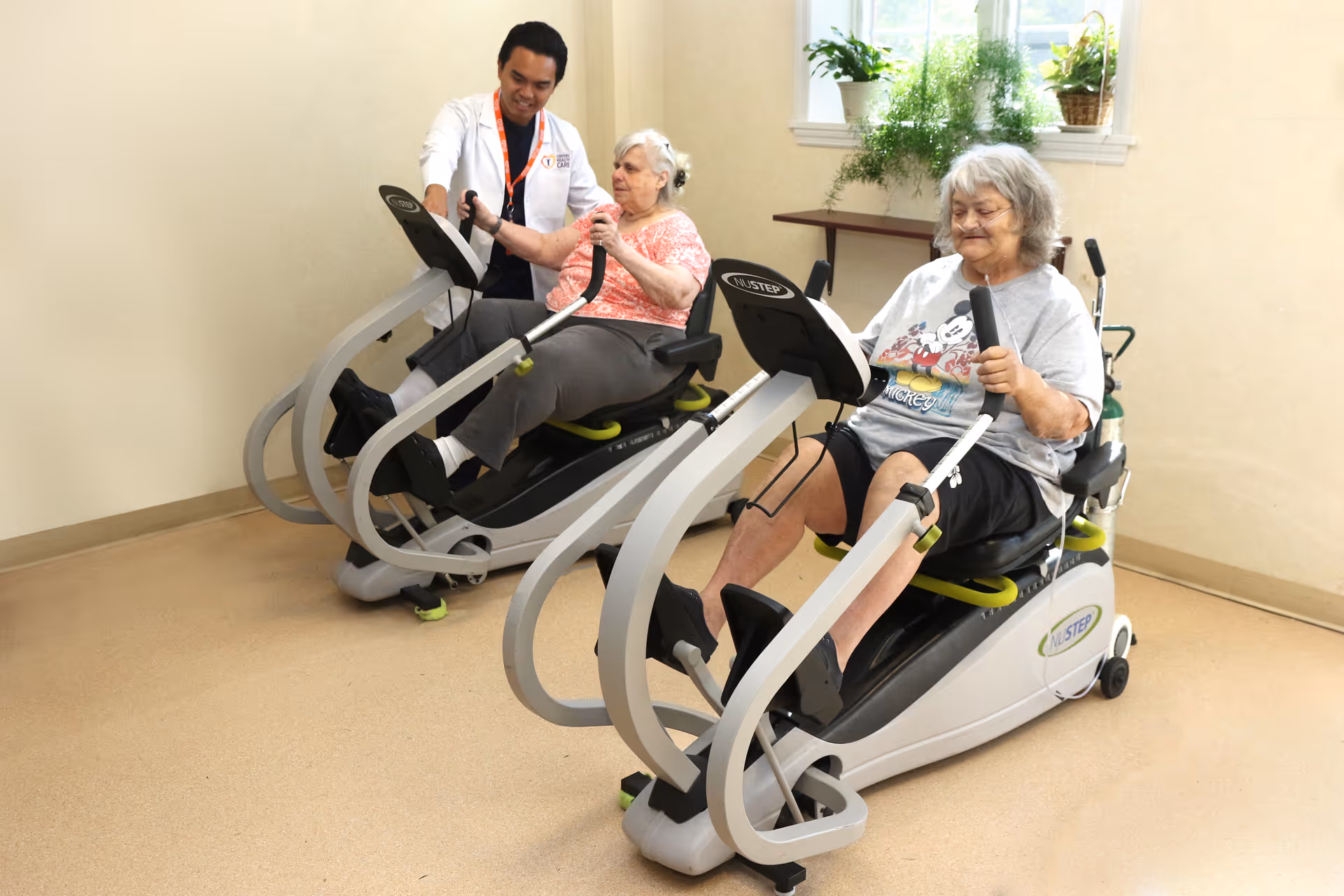 Two elderly women exercising on recumbent cross trainers in a room with beige walls and a window with plants on the windowsill. A healthcare professional in a white coat is assisting one of the women.
