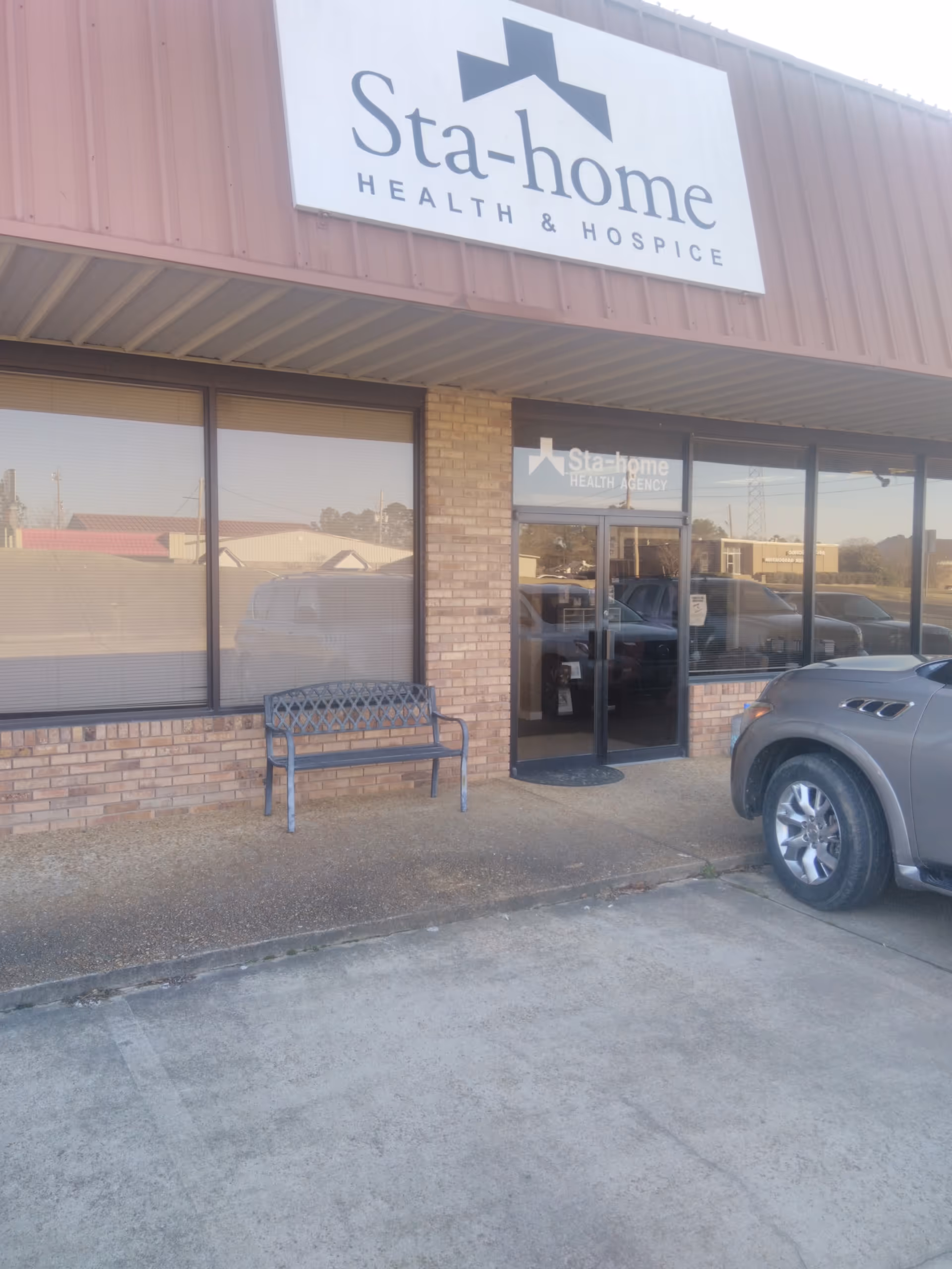 Exterior view of Sta-Home Health Agency storefront with a bench and parked SUV outside.