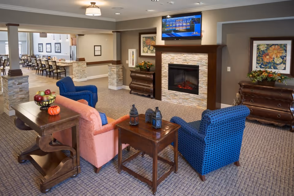 A cozy living room area in a memory care community featuring a pink sofa and two blue armchairs arranged around a wooden coffee table with decorative lanterns. The room has a carpeted floor, a stone fireplace with a TV mounted above it, and two wooden dressers with floral artwork and plants on top. In the background, a dining area with tables and chairs is visible.