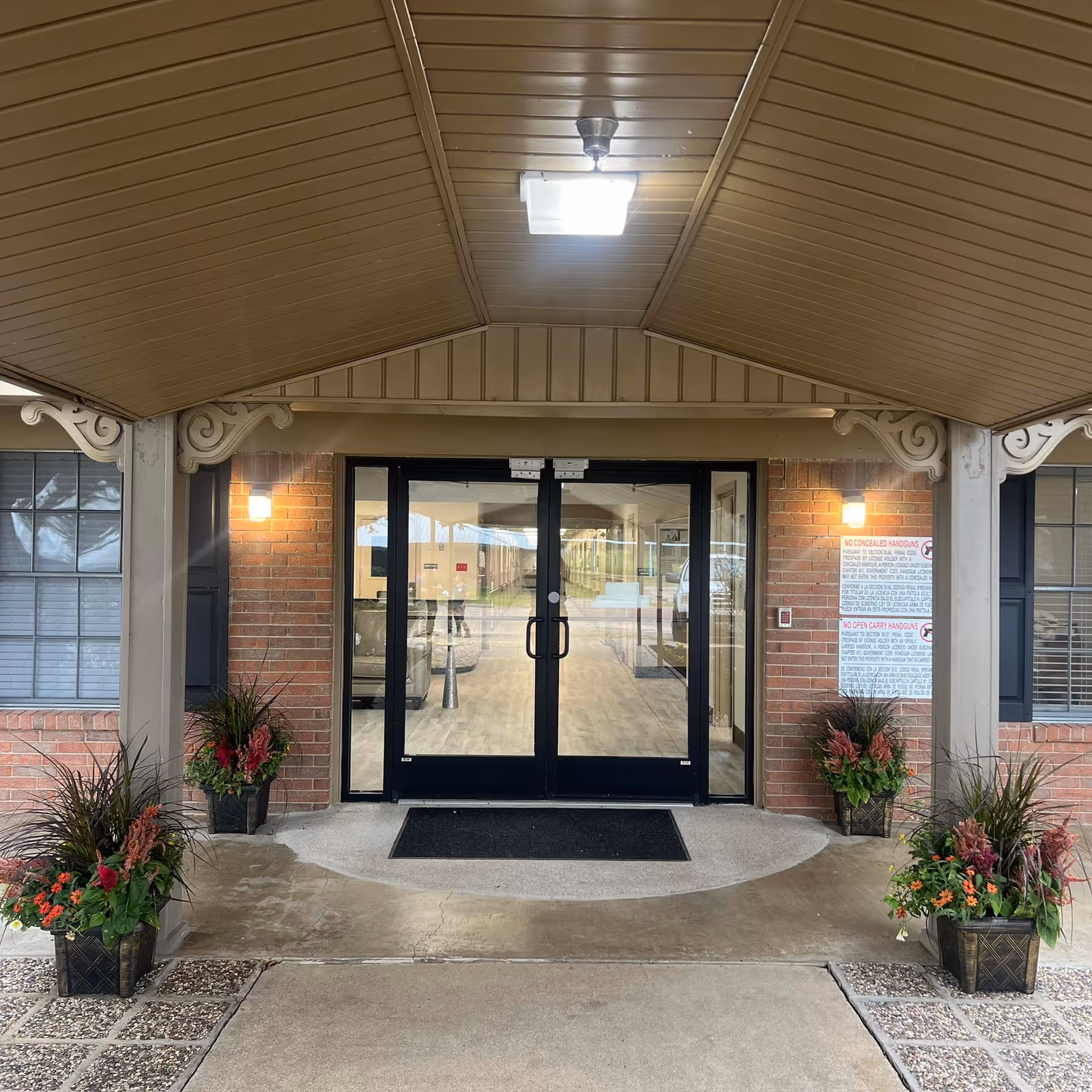 Covered entrance to a building with double glass doors, brick walls, and decorative columns. There are four planters with green and red plants on either side of the entrance. A black doormat is placed in front of the doors, and a light fixture is mounted on the ceiling above the entrance.