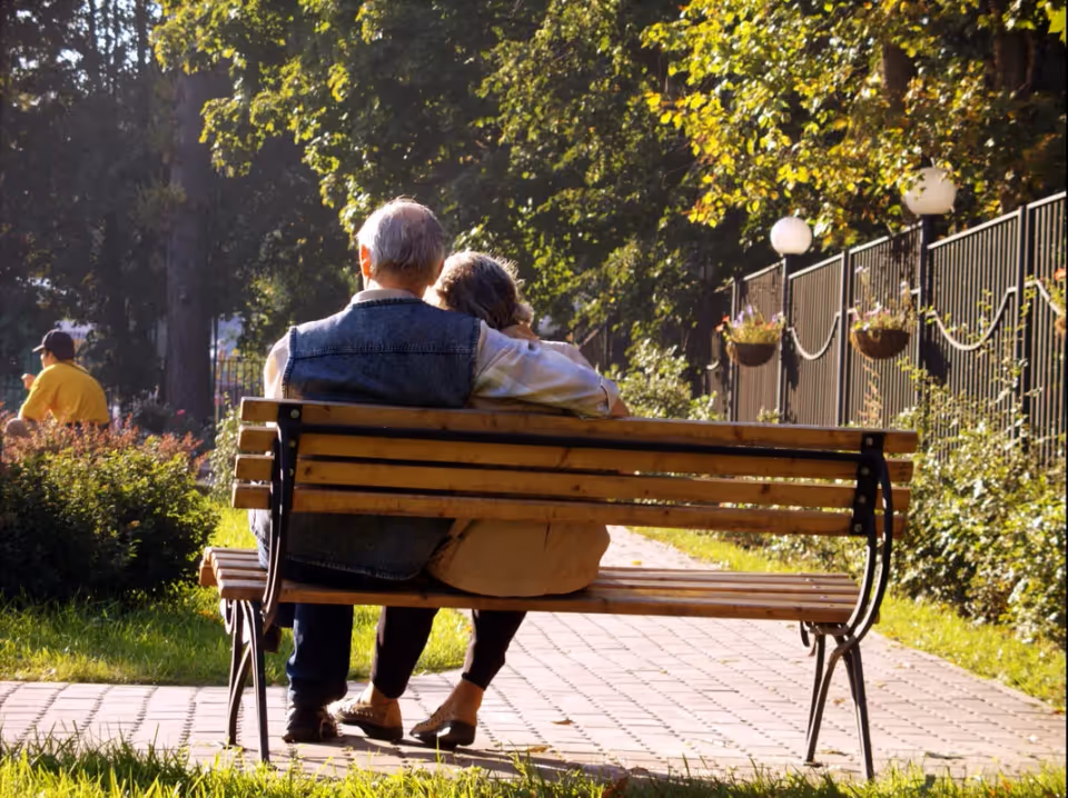An older couple sits closely on a wooden park bench, viewed from behind, enjoying a sunny tree-lined walkway.