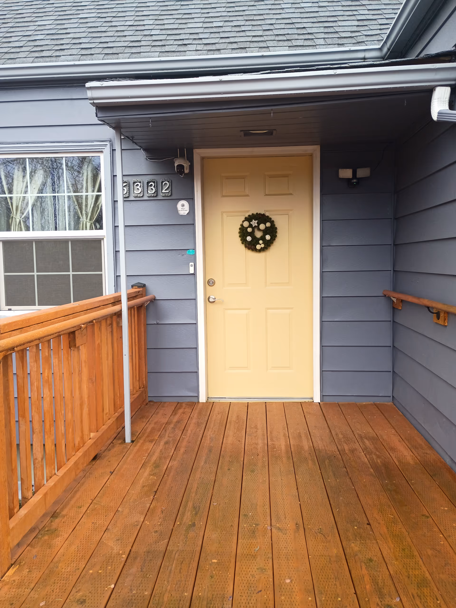 Front entrance of a blue building with a yellow door and wreath on a wooden deck.