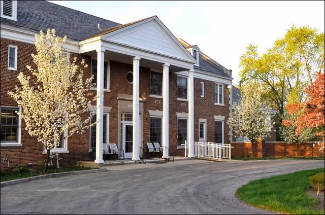 Brick senior living facility entrance with white columns, flowering trees, and a curved driveway.