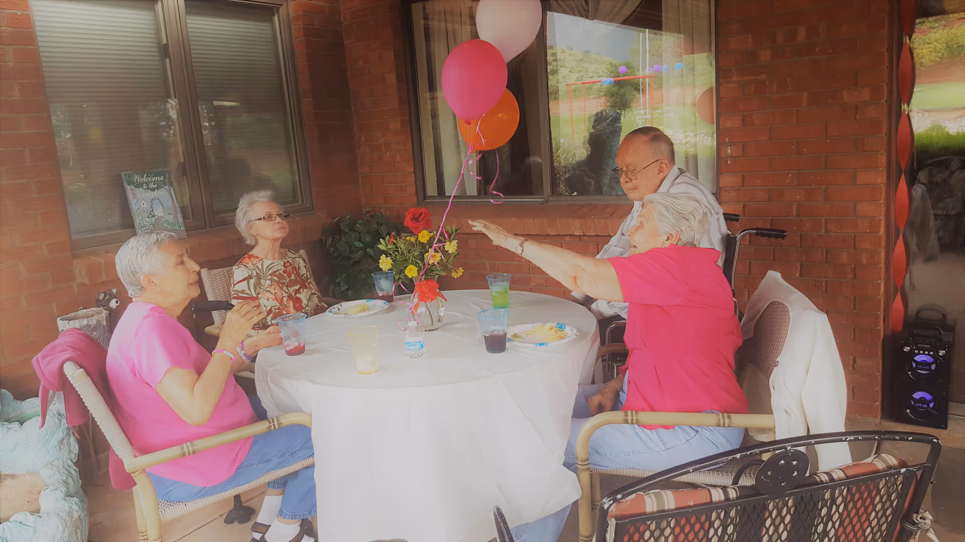 Four elderly people sitting around a round table covered with a white tablecloth, enjoying a meal and drinks on a covered patio with brick walls. The table is decorated with a vase of flowers and colorful balloons. One person is in a wheelchair, and there is a speaker on the floor nearby.