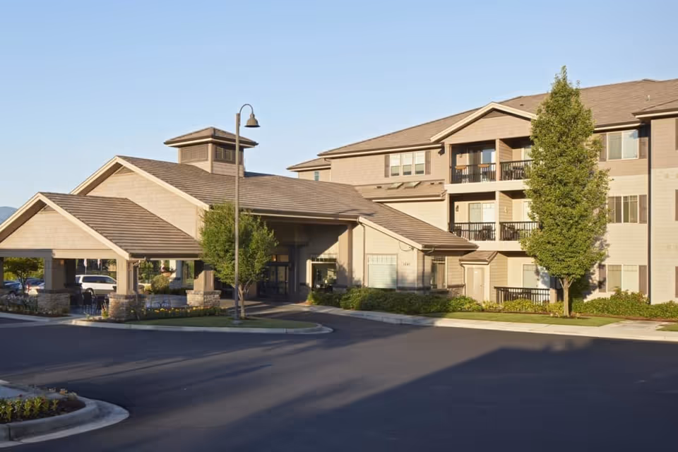 Exterior view of a senior living facility building with a covered entrance, multiple windows, balconies, and surrounding landscaping including trees and shrubs under a clear sky.