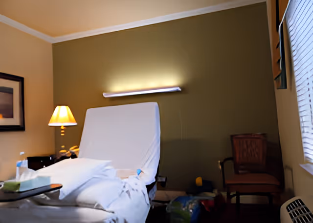 Hospital-style patient room with an adjustable bed, pillows and blanket, a lit bedside lamp, chair, and window blinds.