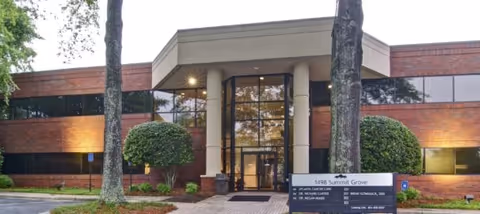 Brick medical building entrance with glass doors, columns, and landscaped shrubs.