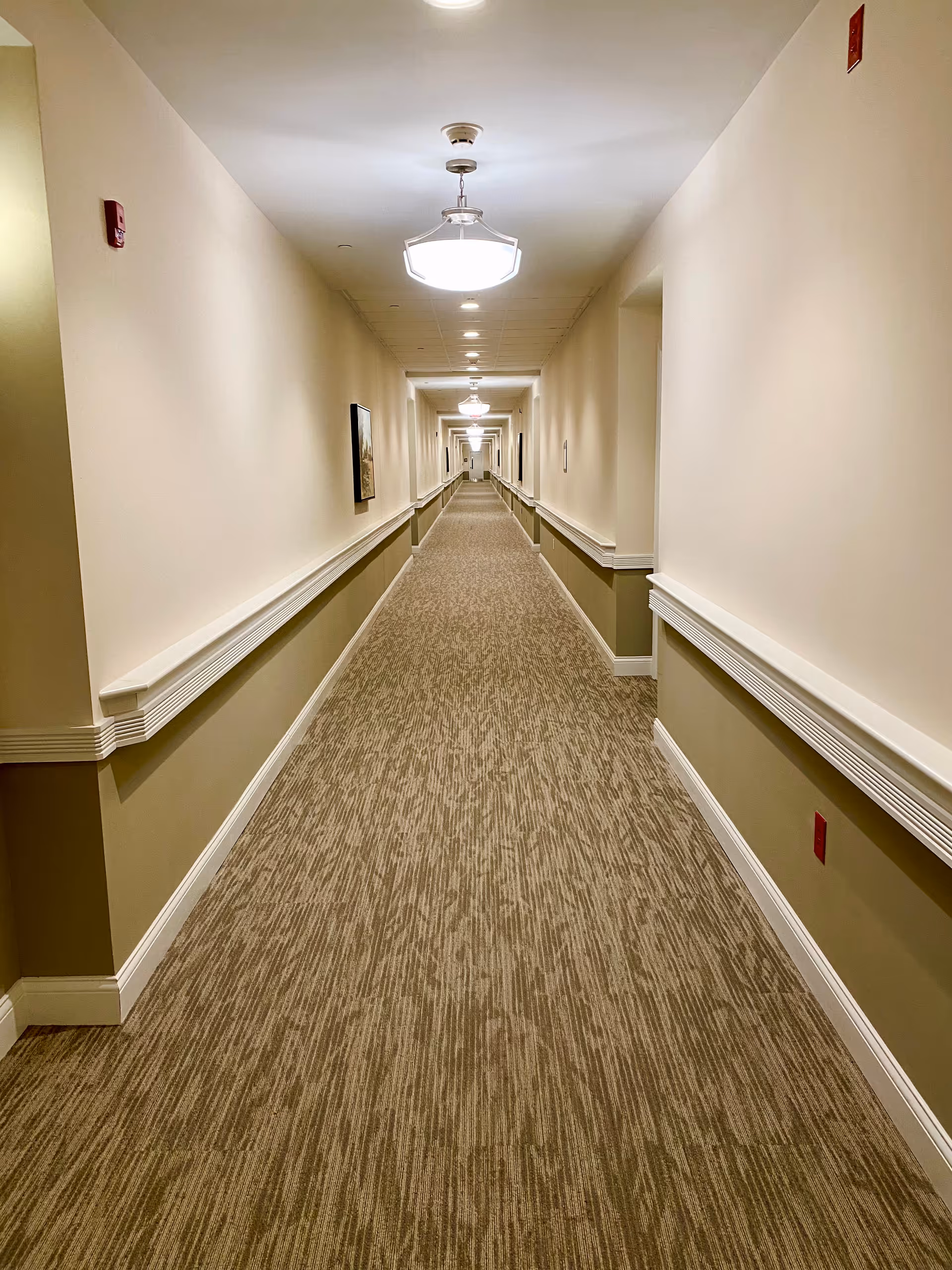 Long beige hallway with ceiling lights and wall railings in a senior living facility.