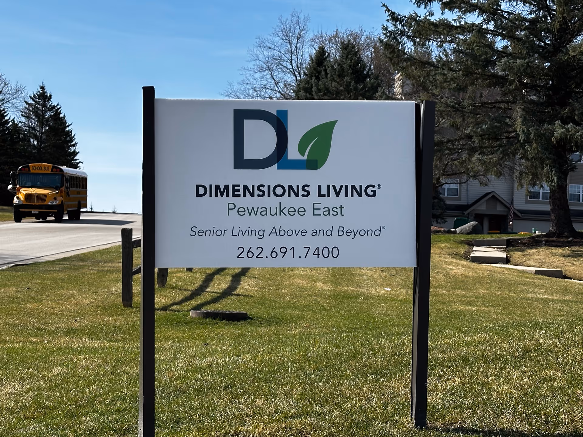 A yard-mounted sign reading "Dimensions Living Pewaukee East" in front of a grassy lawn with the facility and a school bus visible in the background.