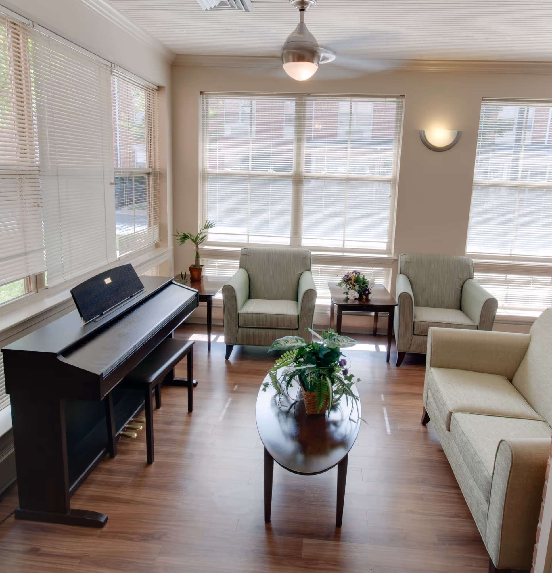 A bright living room with large windows covered by blinds, featuring a ceiling fan with light, a dark wooden piano with a matching bench, two beige armchairs, a beige loveseat, two small side tables with plants and flowers, and a dark wooden coffee table with a green potted plant on it. The floor is wooden.