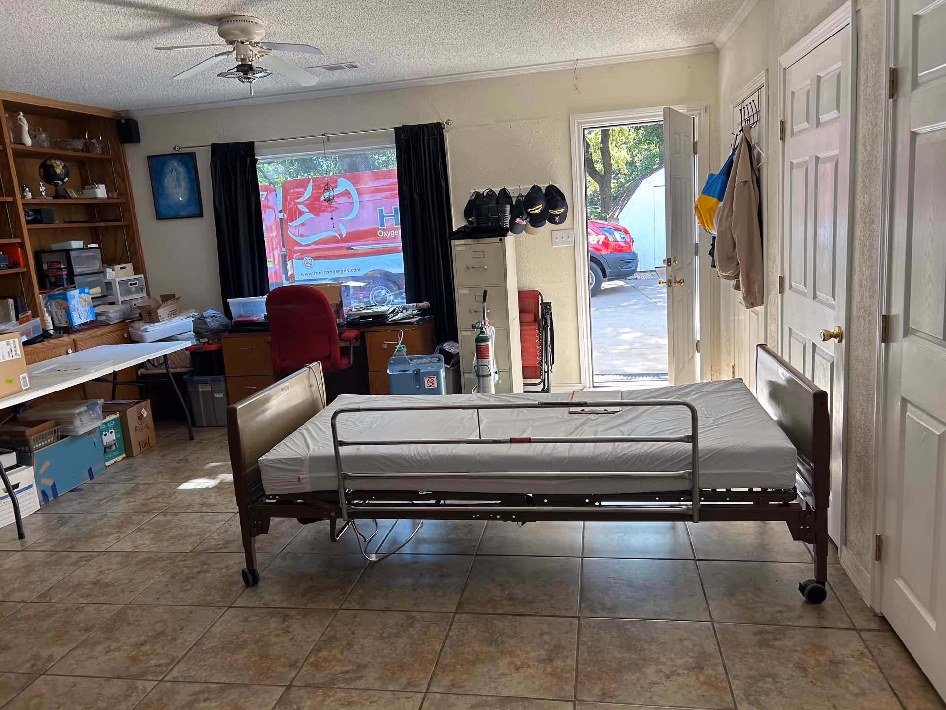 A hospital-style bed in a cluttered resident room with desk and shelves.