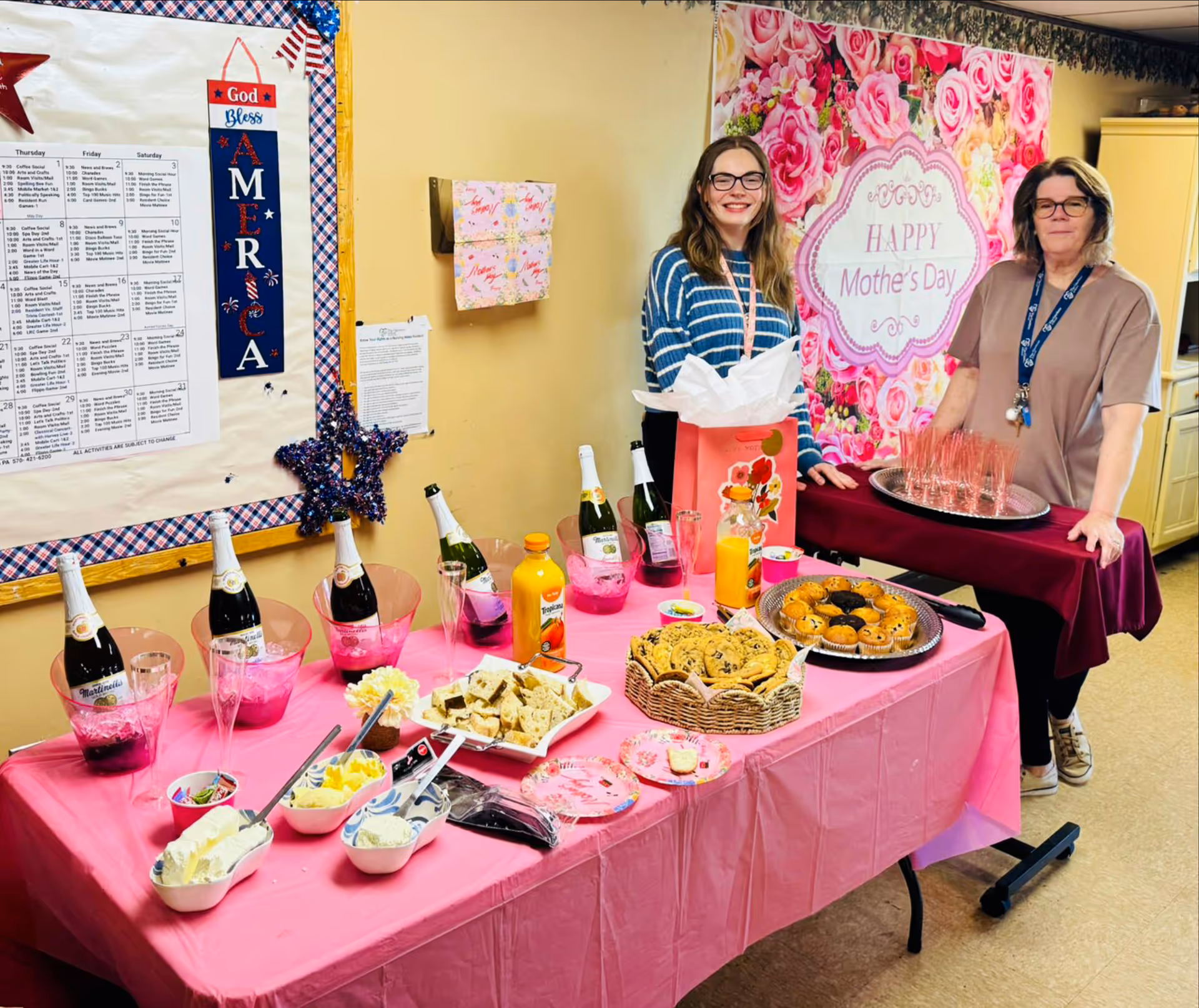 Two women stand behind a pink table laden with drinks, snacks, and a Mother's Day backdrop.