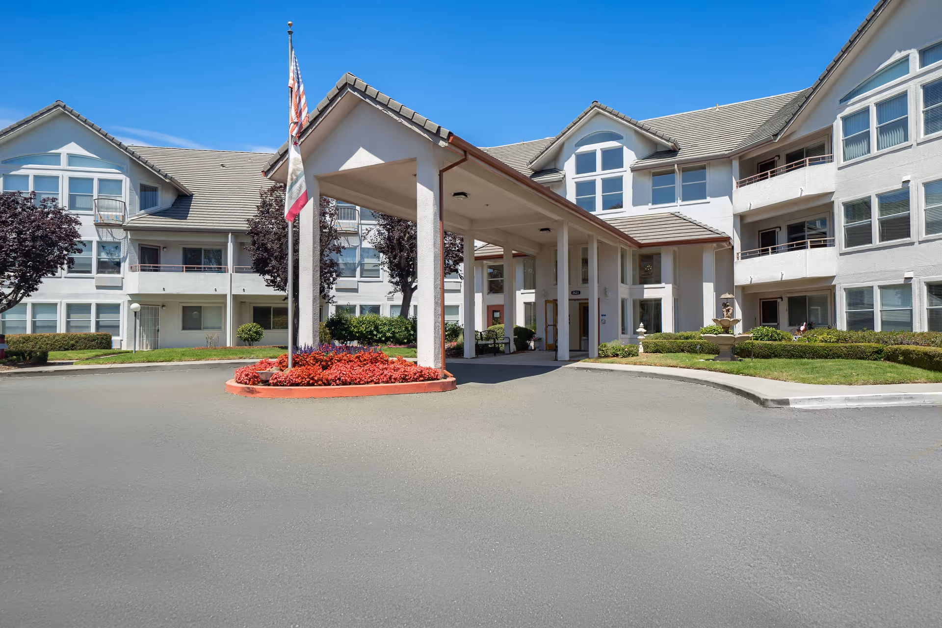 White multi-story senior living building with covered entrance, flagpole, and landscaped roundabout under a blue sky.