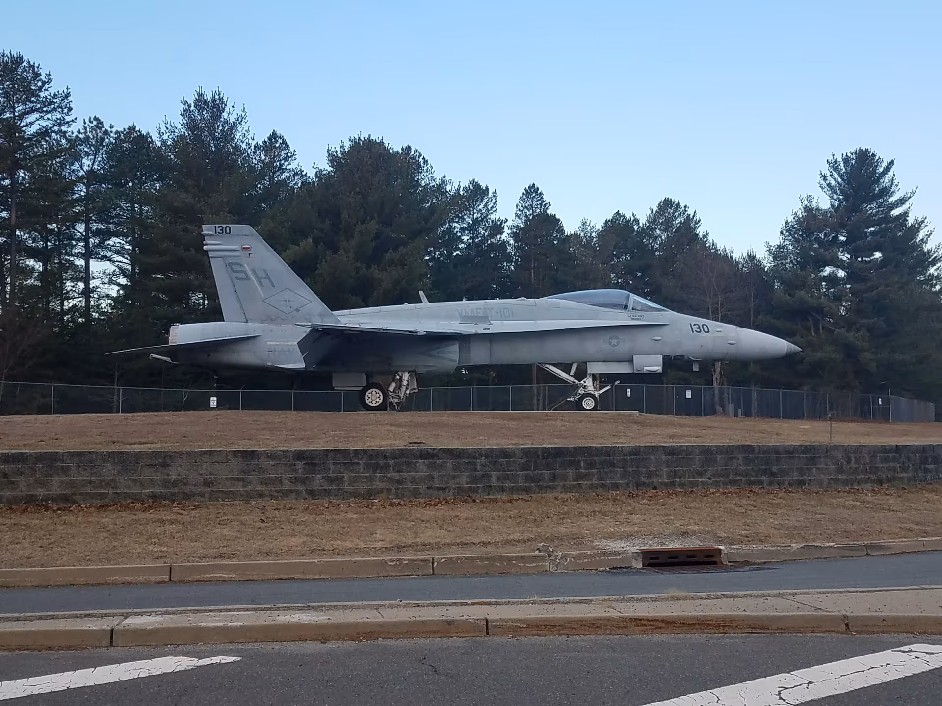 A silver fighter jet on display outdoors behind a chain-link fence.