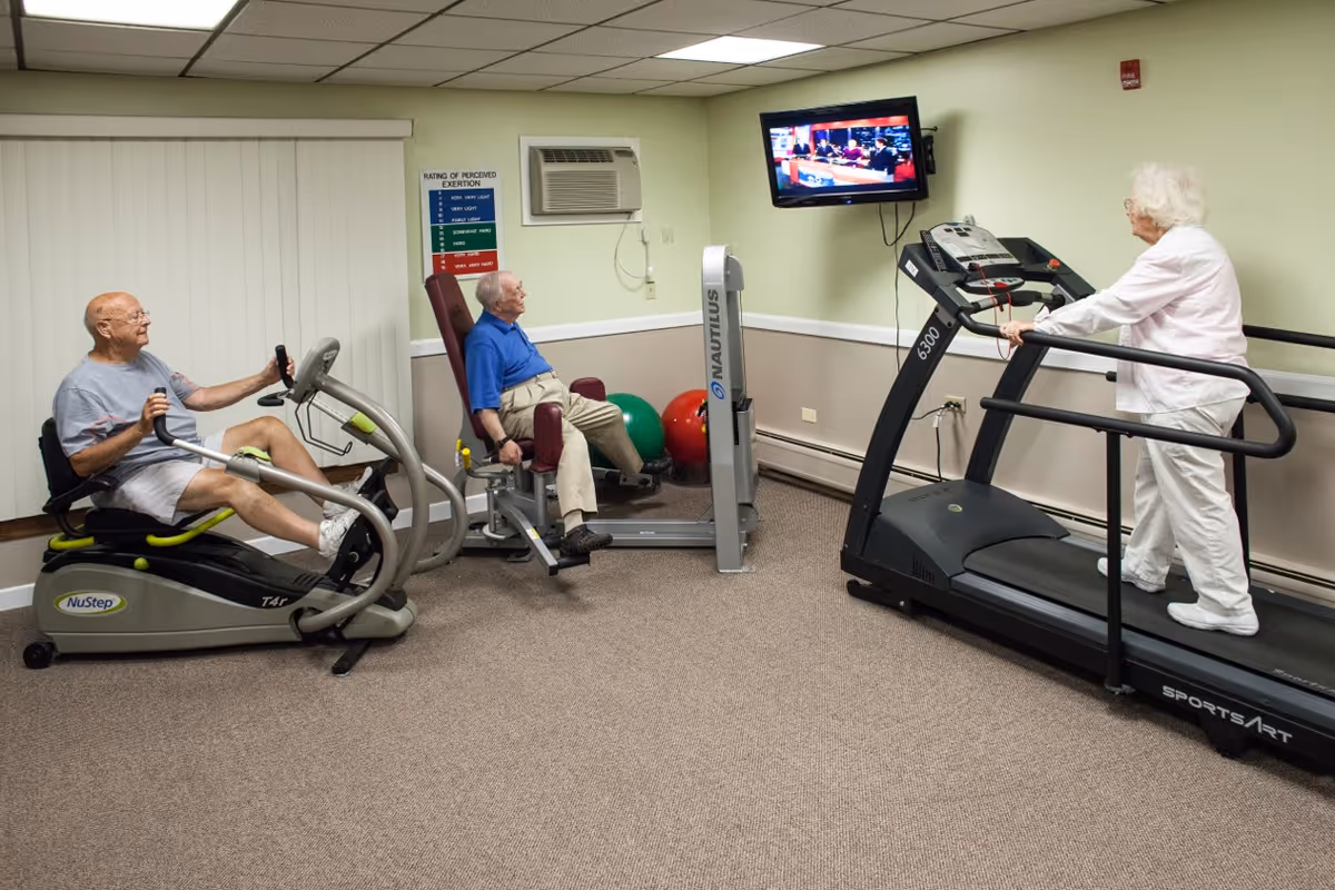 Three elderly residents exercising on machines in a small indoor fitness room with a wall-mounted TV.