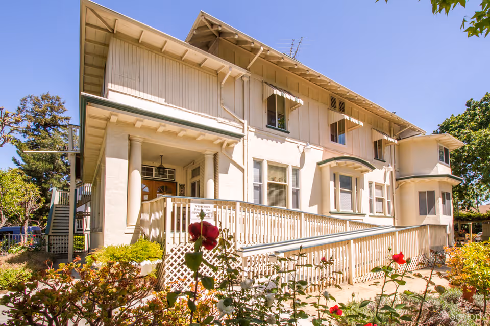 White two-story senior living building with porch, railing, and blooming roses in foreground.