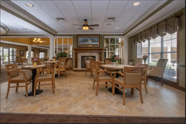 Bright communal dining and lounge area with round tables and chairs arranged around a fireplace and large windows.
