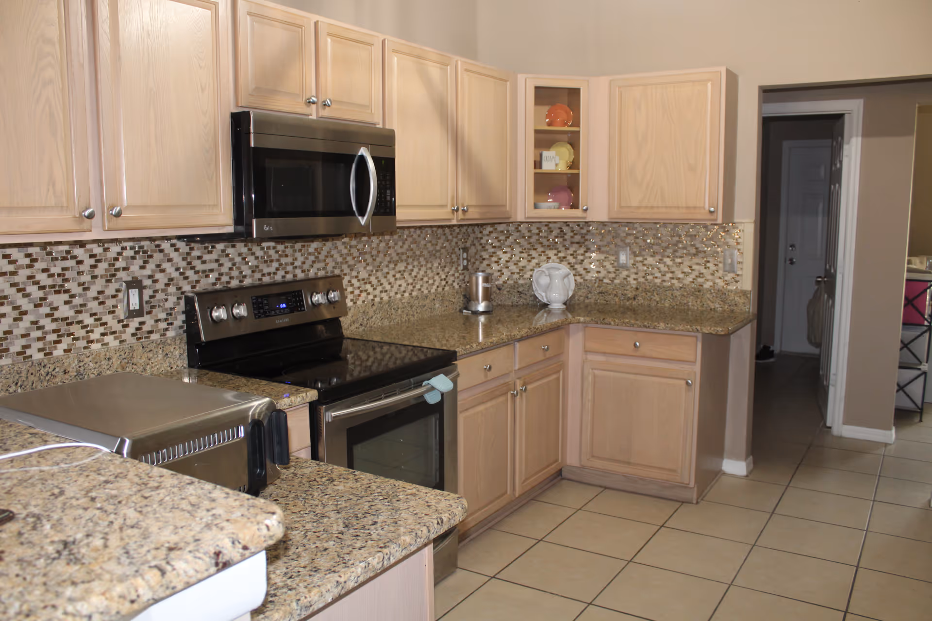A kitchen with light wood cabinets, granite countertops, and a mosaic tile backsplash. The kitchen features a stainless steel microwave above a black electric stove and oven. There is a toaster oven on the counter and a small decorative plate display in a glass-front cabinet. The floor is tiled and there is an open doorway leading to another room.