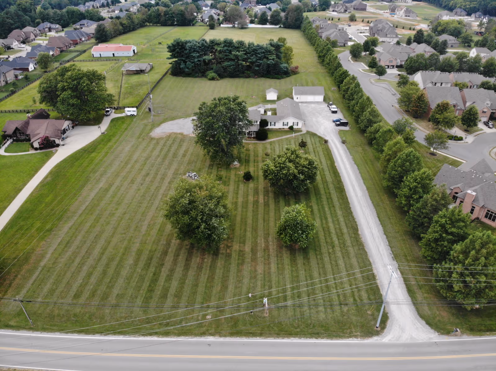Aerial view of a large grassy property with a few trees and a single-story house near the center. The property is bordered by a gravel driveway leading to a road, with neighboring houses and residential streets visible in the background.