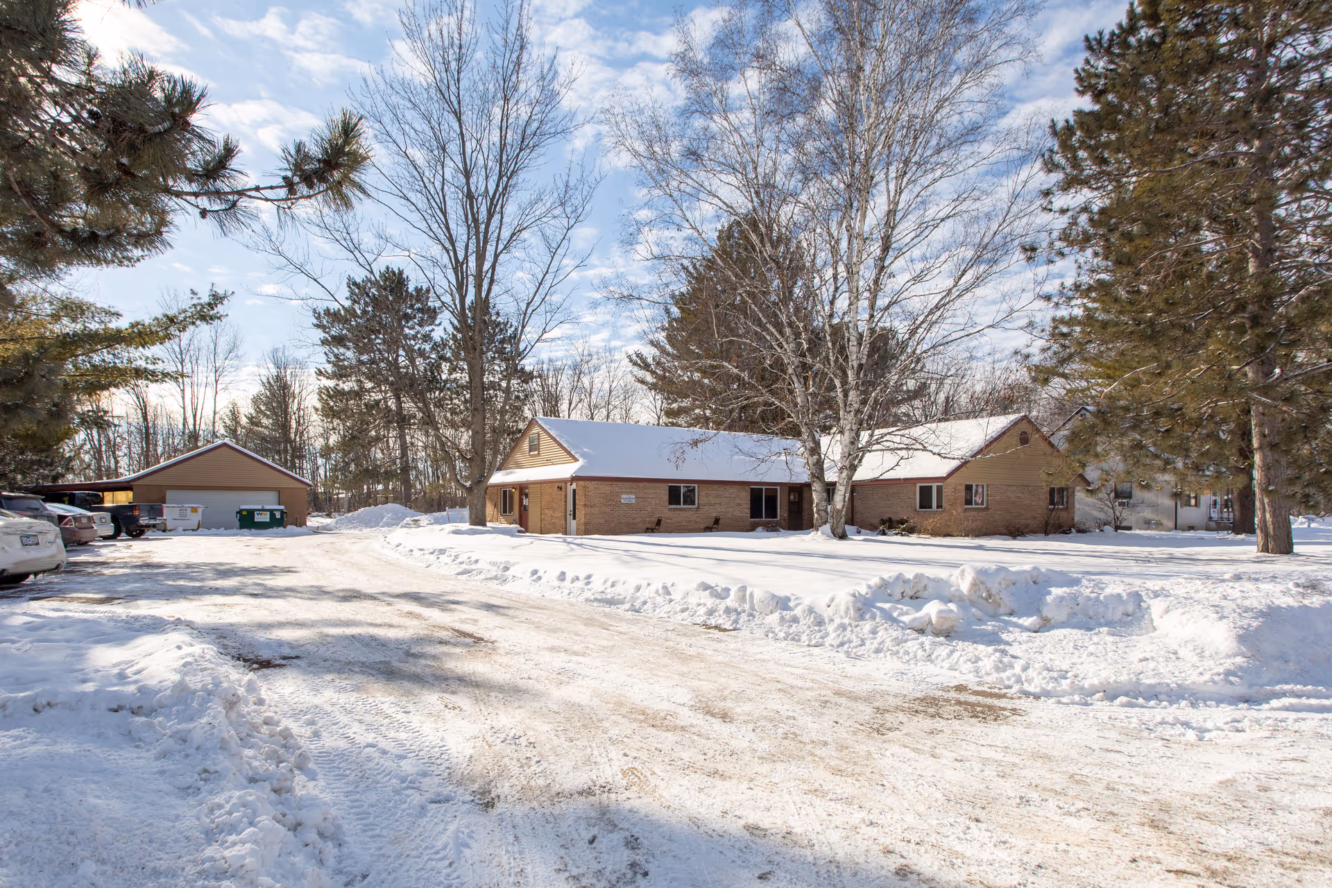 Snow-covered VitaCare Living of Mora exterior: a low brick building with surrounding trees.