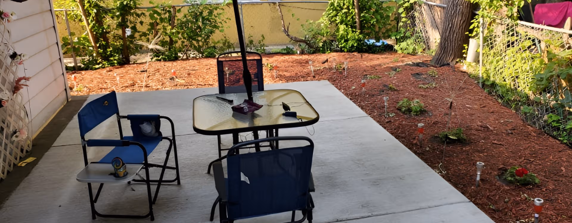Outdoor patio with blue chairs around a glass-top table in a fenced garden.