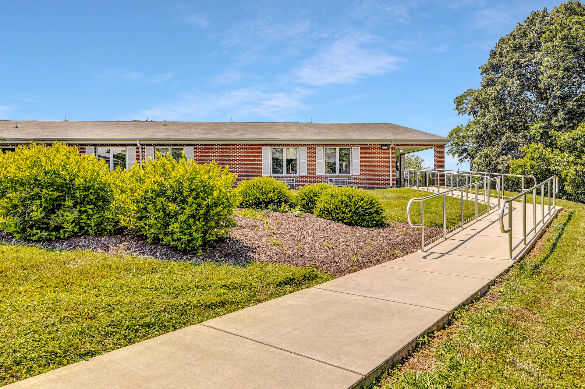 Exterior view of a single-story brick building with white window shutters, surrounded by green bushes and grass. A concrete sidewalk with metal handrails leads up to the entrance on a sunny day with a clear blue sky.