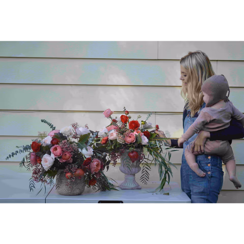 A woman holds a baby beside a table with pink and red flowers outdoors.
