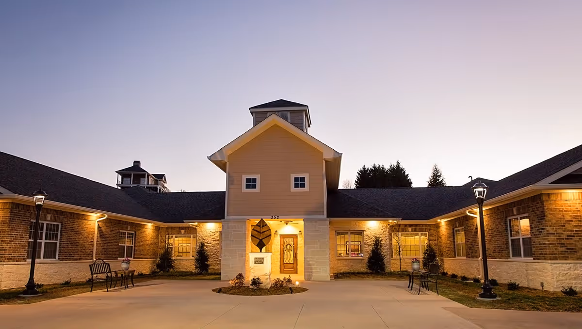 Exterior view of a single-story brick and stone building at dusk with warm lighting. The building has a central entrance with a decorative leaf sculpture on a pedestal in front. There are benches and lamp posts on either side of the entrance pathway.