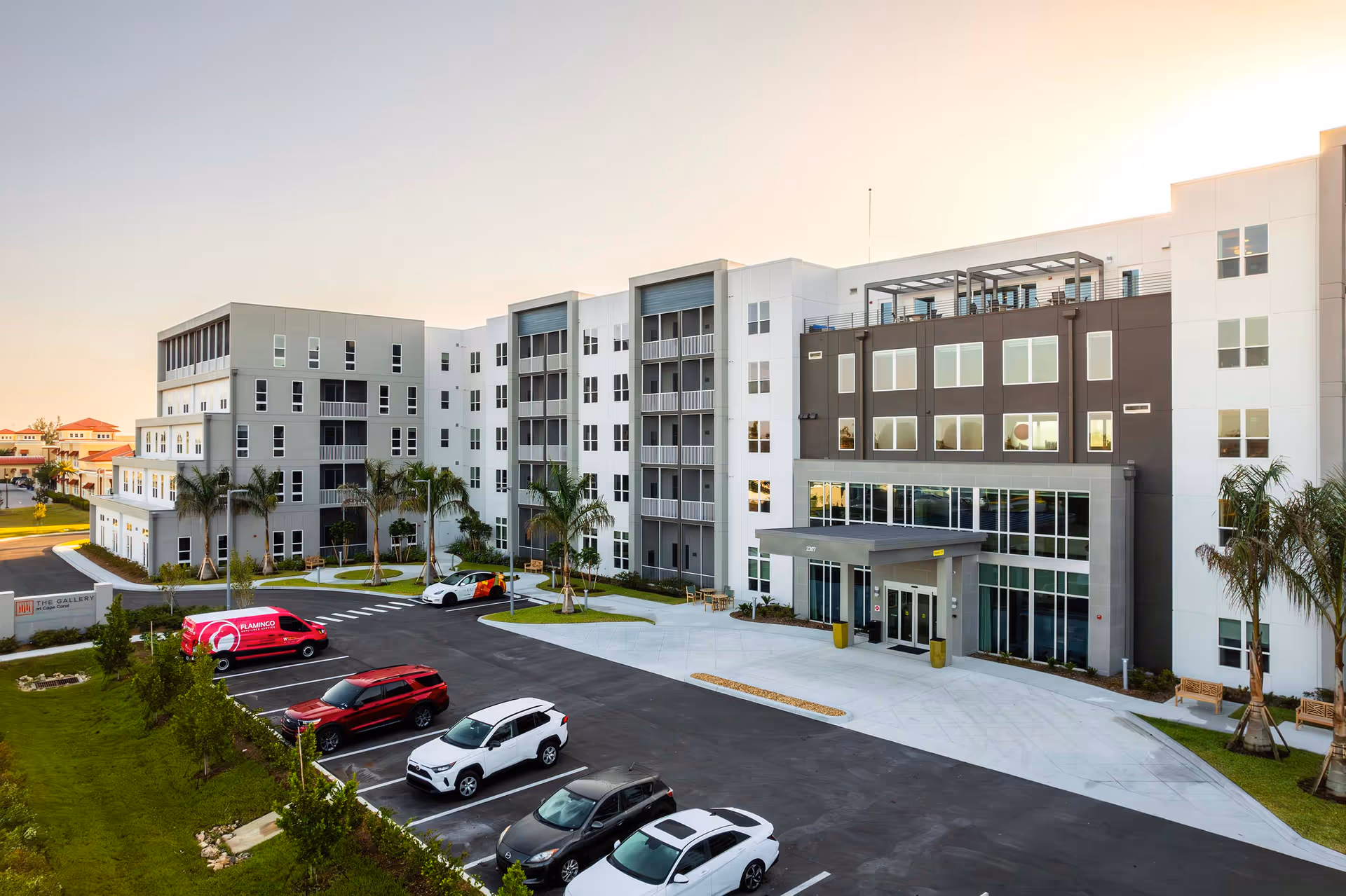 Exterior view of The Gallery at Cape Coral, a modern multi-story senior living facility with a parking lot in front, several parked cars, palm trees, and a clear sky at sunset.