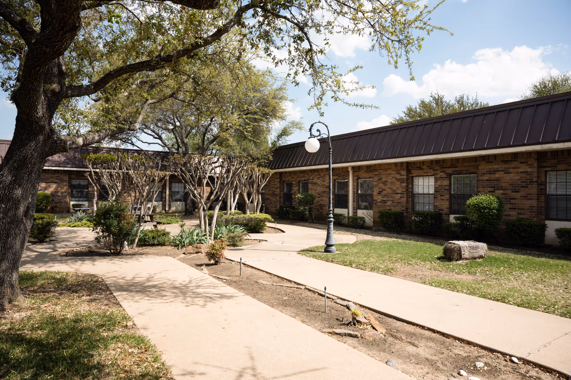 Outdoor courtyard area at Park Bend Rehabilitation & Healthcare Center featuring paved walkways, trimmed trees, bushes, a vintage-style lamp post, and a brick building with multiple windows in the background under a partly cloudy sky.