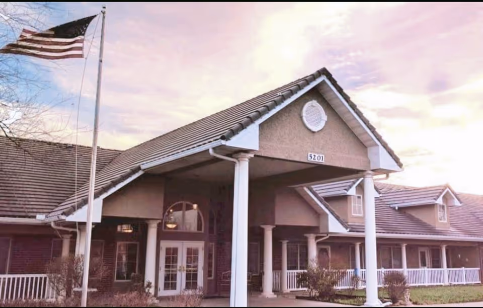 Front entrance of a senior living building with white columns, a covered portico, an American flag, and the address number 5201 visible.